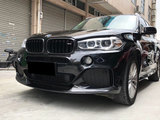 A black BMW SUV is parked on a concrete surface, its headlights and sleek grille visible. Nearby, an urban setting includes closed shutters and a bicycle.