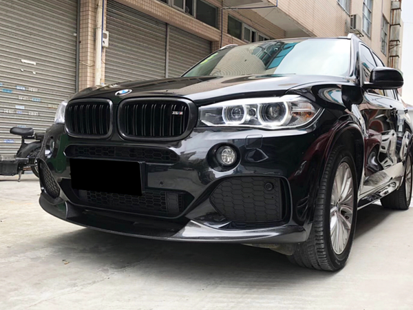 A black BMW SUV is parked on a concrete surface, its headlights and sleek grille visible. Nearby, an urban setting includes closed shutters and a bicycle.