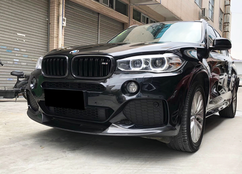A black BMW SUV is parked on a concrete surface, its headlights and sleek grille visible. Nearby, an urban setting includes closed shutters and a bicycle.