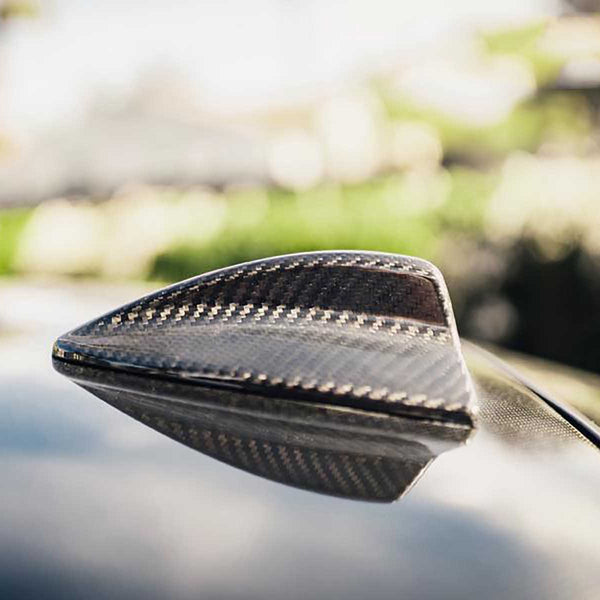Shark fin antenna, made of carbon fiber, sits on a car's roof. It's in a sunlit outdoor environment with blurred greenery in the background.