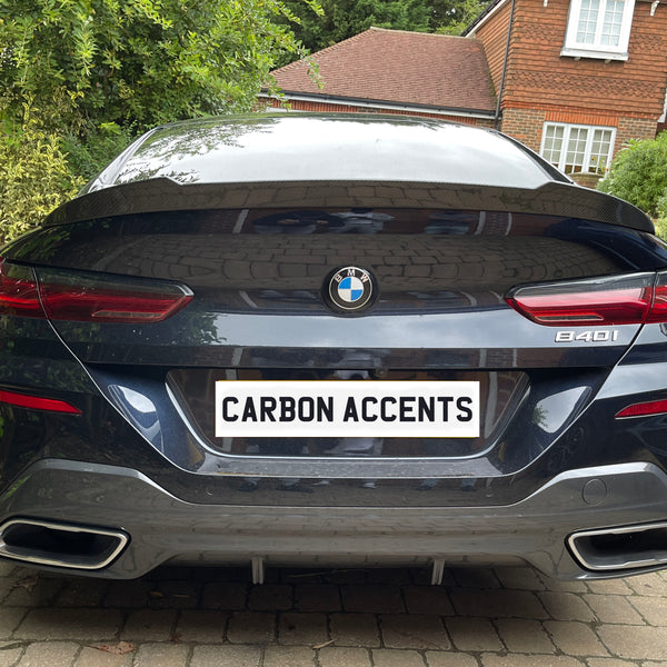 The car, a BMW 840i, is stationary with a license plate reading "CARBON ACCENTS," parked in a driveway surrounded by green bushes and a brick house in the background.