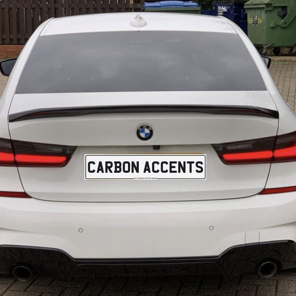 A white BMW parked on a paved surface, featuring rear carbon accents. The context includes a wooden fence and two trash bins. License plate reads: "CARBON ACCENTS.”