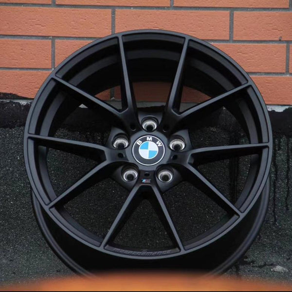 A black BMW car wheel rim with a logo at the center is leaning against a brick wall on a textured ground surface.