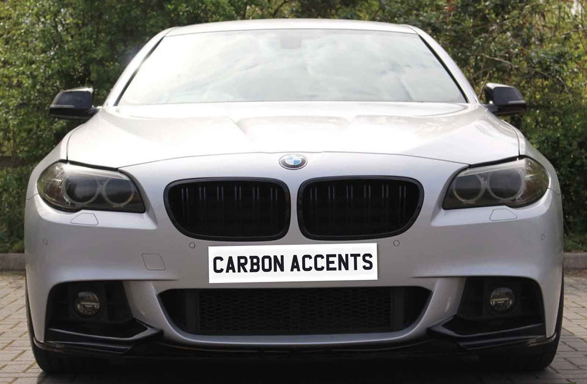 A silver car with a prominent front grille and headlights, parked on a paved area, displays the text "CARBON ACCENTS" on its license plate. Trees are visible in the background.