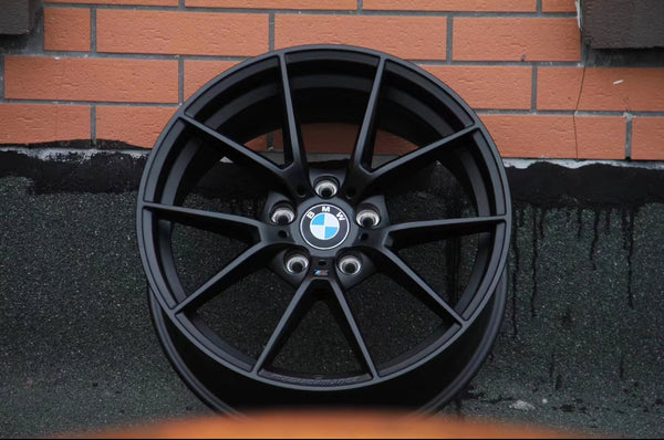 A black BMW alloy wheel rests against a brick wall, featuring the BMW logo in the center and surrounded by asphalt paving.