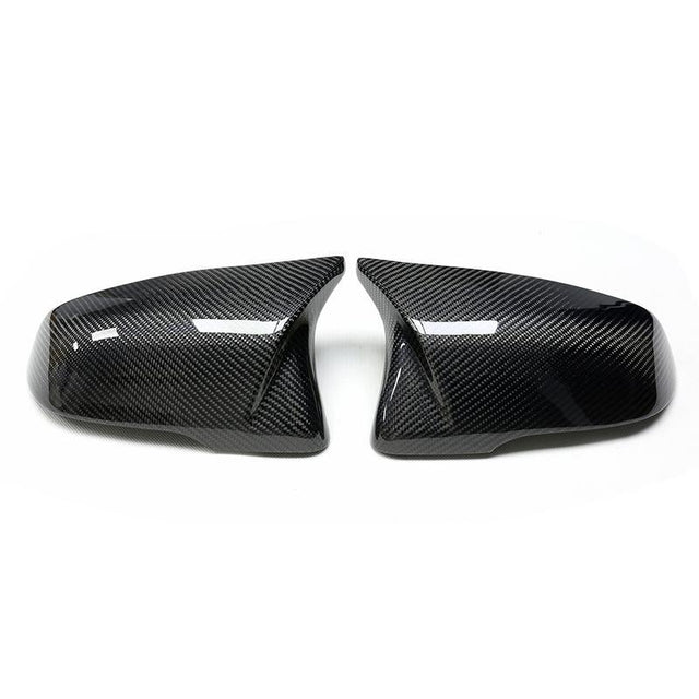 Two glossy carbon fiber car mirror covers are positioned side by side against a plain white background, reflecting light on their textured surface.