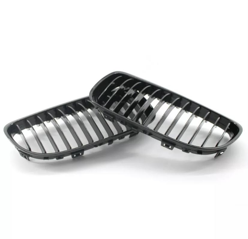 Two black car grilles rest on a white surface, showcasing their shiny, vertical slats and curved rectangular frames, designed for vehicle front-end placement.