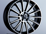 A silver and black alloy car wheel stands upright on a black pedestal. The central hub features the Mercedes-Benz logo. The background is a plain, light gray surface.