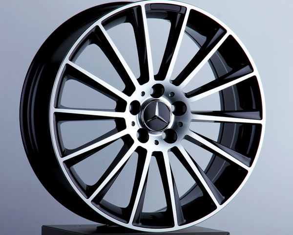 A silver and black alloy car wheel stands upright on a black pedestal. The central hub features the Mercedes-Benz logo. The background is a plain, light gray surface.