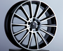 A silver and black alloy car wheel stands upright on a black pedestal. The central hub features the Mercedes-Benz logo. The background is a plain, light gray surface.