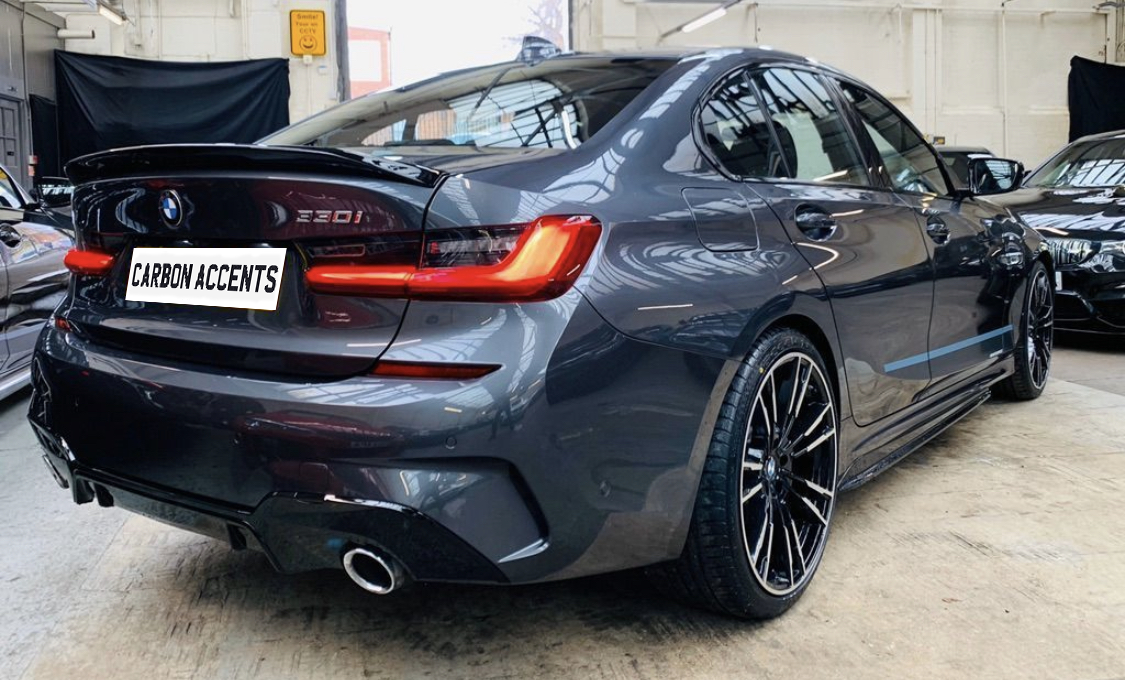 Car parked indoors, featuring a sleek gray BMW 320i with visible dual exhausts and tinted windows. The license plate reads "CARBON ACCENTS." The setting is an industrial garage.