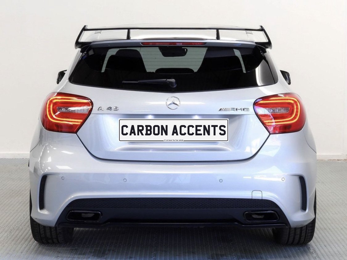 A silver hatchback car, viewed from the rear, has a prominent spoiler and dual exhaust. Text reads "CARBON ACCENTS" on the license plate. It is indoors on a patterned floor.