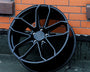 A black alloy wheel stands upright against an orange brick wall on a gray concrete surface. The wheel features multiple spokes and a central hub surrounded by five lug holes.