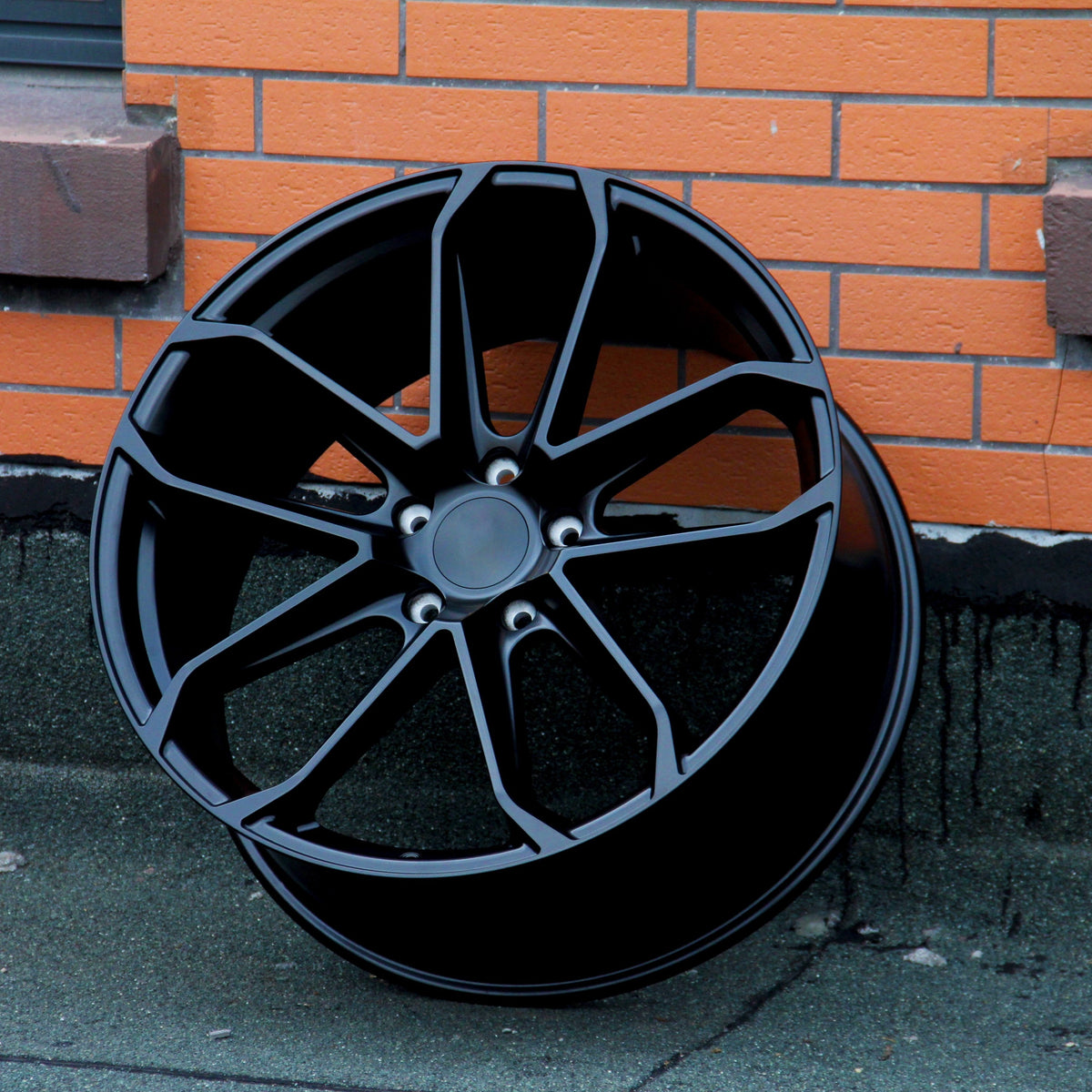 A black alloy wheel stands upright against an orange brick wall on a gray concrete surface. The wheel features multiple spokes and a central hub surrounded by five lug holes.