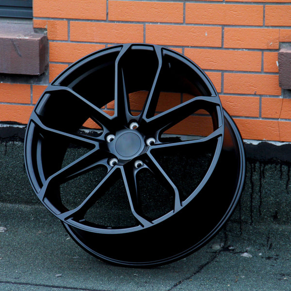 A black alloy wheel stands upright against an orange brick wall on a gray concrete surface. The wheel features multiple spokes and a central hub surrounded by five lug holes.