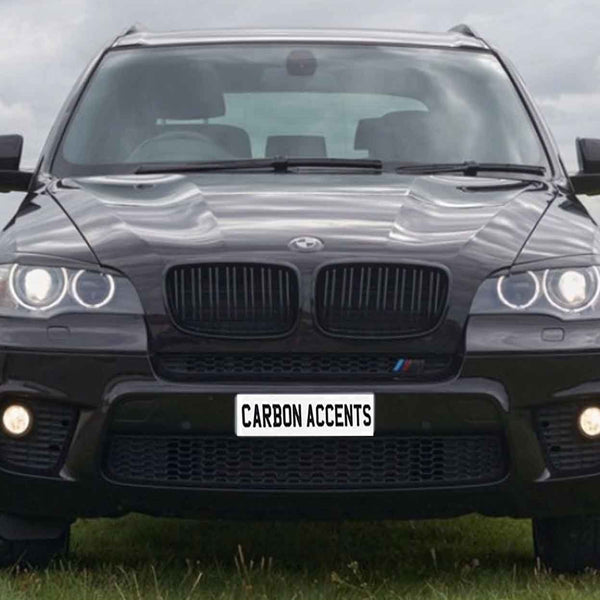 A black SUV with illuminated headlights is parked on grass, appearing robust and sleek. The license plate reads "CARBON ACCENTS." Behind is a cloudy sky and a distant tree line.
