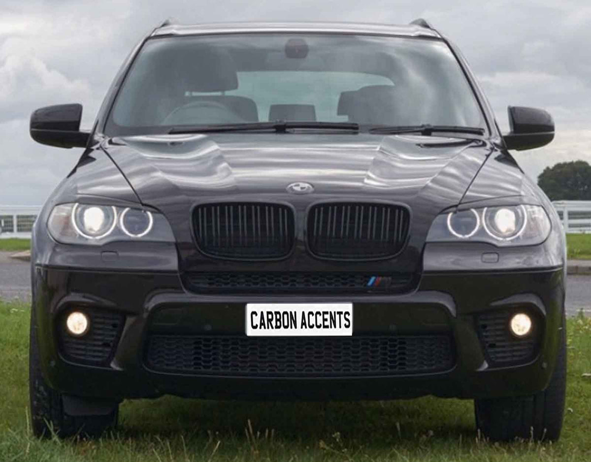 A black SUV with illuminated headlights is parked on grass, appearing robust and sleek. The license plate reads "CARBON ACCENTS." Behind is a cloudy sky and a distant tree line.