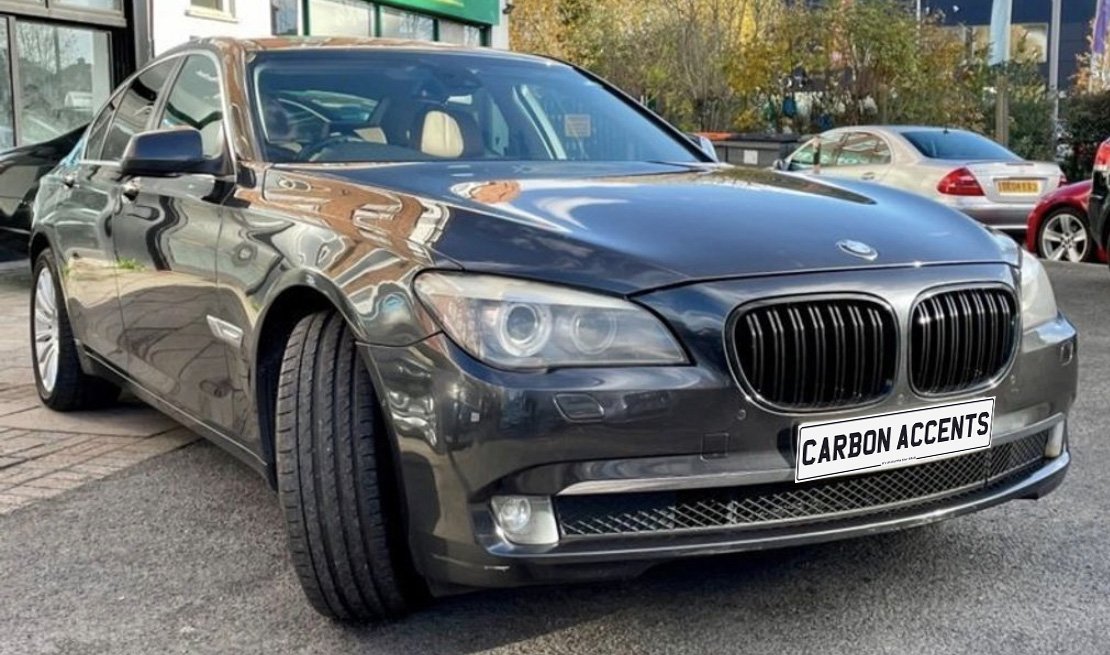 A sleek, dark-colored car with the front wheel turned slightly to the side is parked on a street. The license plate reads "CARBON ACCENTS," surrounded by other parked vehicles.