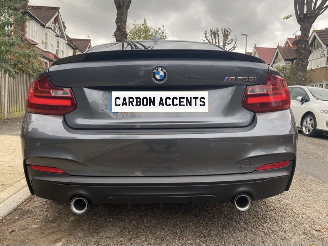 A gray BMW M235i, with dual exhausts, is parked on a residential street. The license plate reads "CARBON ACCENTS," and houses and trees line the background.