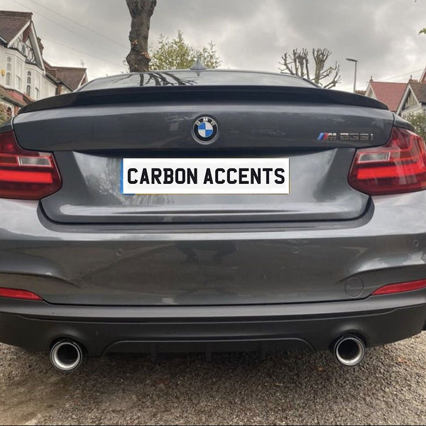 A gray BMW M235i, with dual exhausts, is parked on a residential street. The license plate reads "CARBON ACCENTS," and houses and trees line the background.