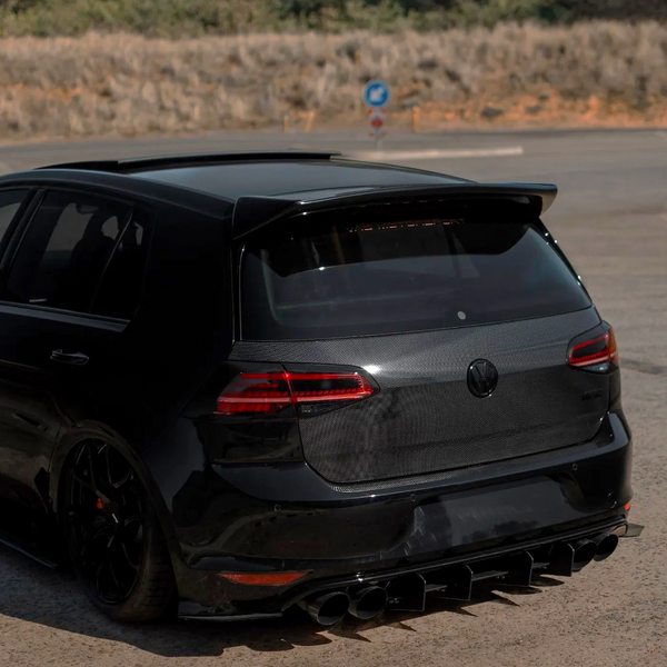 A black hatchback car is parked on a pavement with a carbon fiber rear, featuring prominent tailpipes and a roof spoiler, surrounded by a dry, grassy landscape.