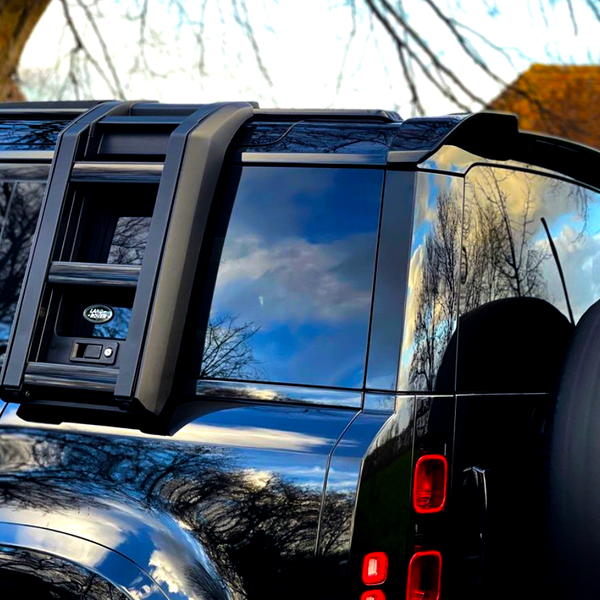 A black SUV with a ladder mounted on the rear window is parked outdoors in a grassy area with trees nearby. Reflections of clouds and trees are visible on the windows.