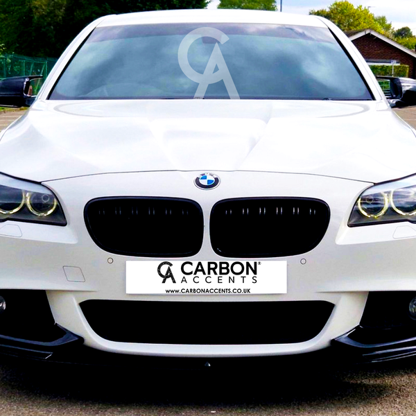 A white BMW sedan is parked, facing forward, under clear skies in a parking lot with greenery and buildings. Text on the front reads: "CARBON ACCENTS www.CARBONACCENTS.CO.UK".