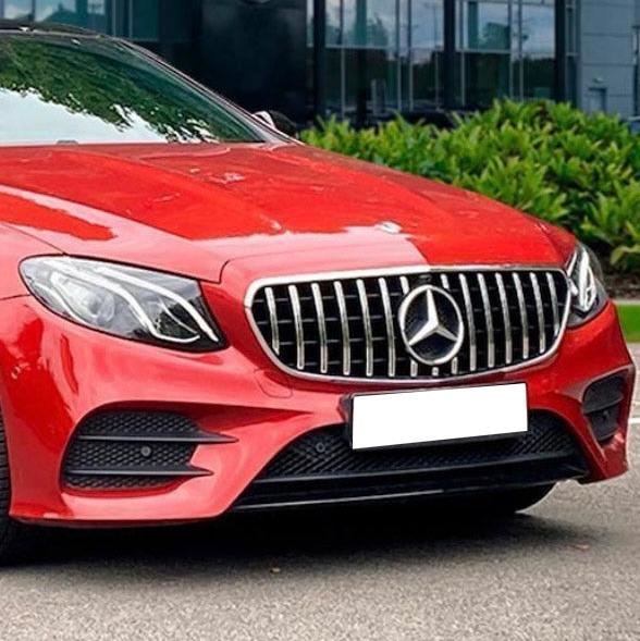 A red Mercedes-Benz car with a prominent grille and headlights is parked on a street, with lush green bushes and a glass building in the background.
