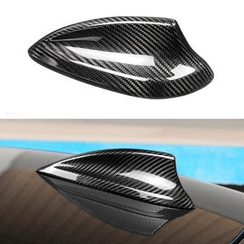 Carbon fiber car antenna cover, sleekly curved, resting on a vehicle roof. The background shows a blurred blue and tan environment, indicating an outdoor setting.