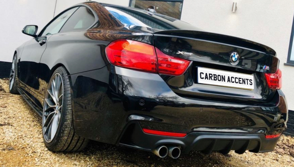 A black sports car with dual exhaust is parked on a gravel driveway beside a white building. The license plate reads "CARBON ACCENTS."