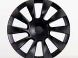A black, multi-spoke car wheel cover with a subtle central logo sits upright against a plain white background, showcasing its sleek, aerodynamic design.