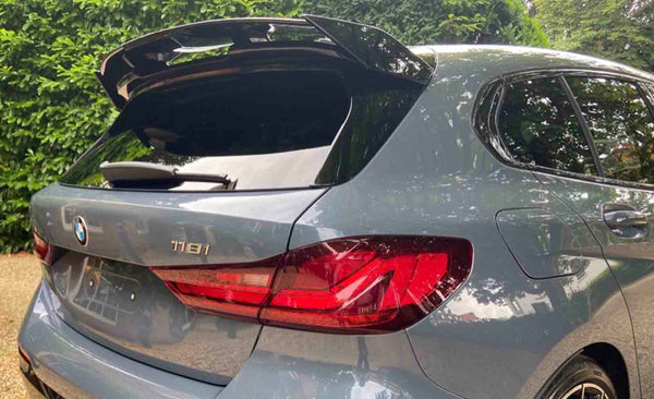 A gray BMW 118i hatchback with an open trunk is parked on a gravel driveway, surrounded by greenery, under a partly cloudy sky.