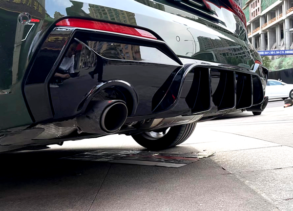 Car exhaust pipe releasing emission, surrounded by a glossy black rear bumper with vents. Reflective cityscape in the metal surface, parked on an urban street.