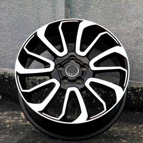 A black and silver alloy wheel rests against a textured gray wall, standing on rough concrete, creating an urban, industrial setting.