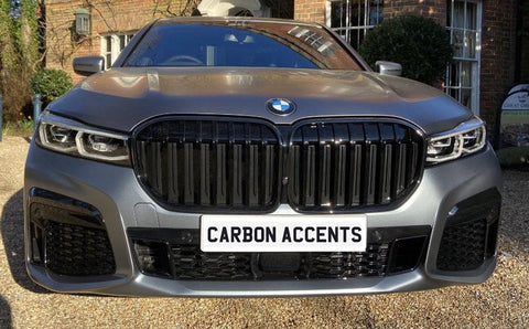 A BMW car parked on a gravel driveway features a prominent grille. The number plate reads "CARBON ACCENTS." A brick building with greenery is in the background.
