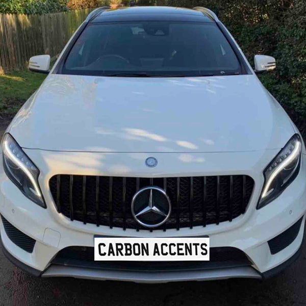 A white SUV with a prominent front grille and headlights parked on a driveway. The license plate reads "CARBON ACCENTS." Surrounded by a wooden fence and greenery.