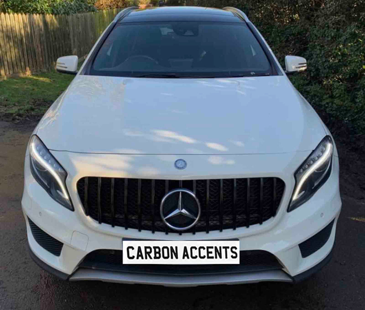 A white SUV with a prominent front grille and headlights parked on a driveway. The license plate reads "CARBON ACCENTS." Surrounded by a wooden fence and greenery.