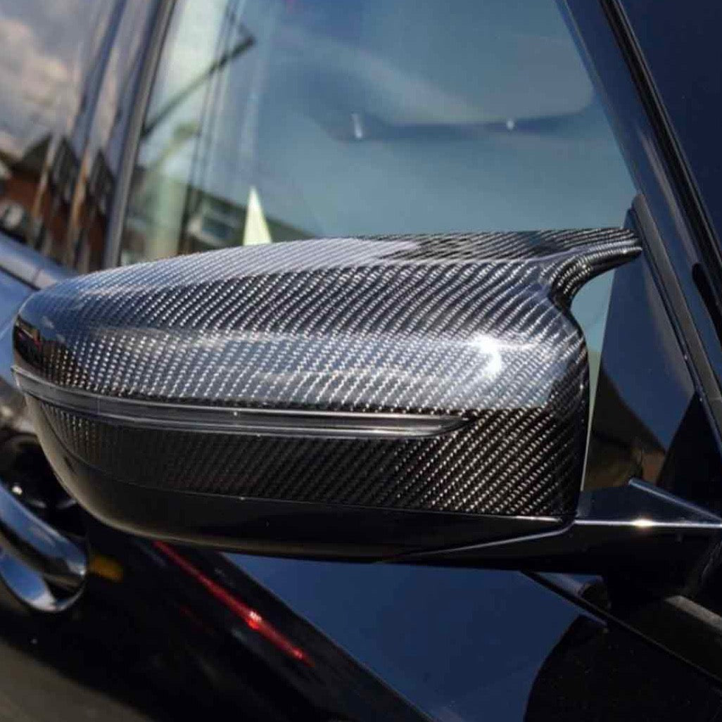 A carbon fiber car side mirror, reflecting light, is attached to the sleek black exterior of a vehicle, with a blurred urban background visible.