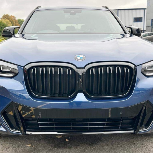 A blue BMW SUV is parked in a parking lot, displaying a prominent front grille and headlights, with trees and industrial buildings in the background.