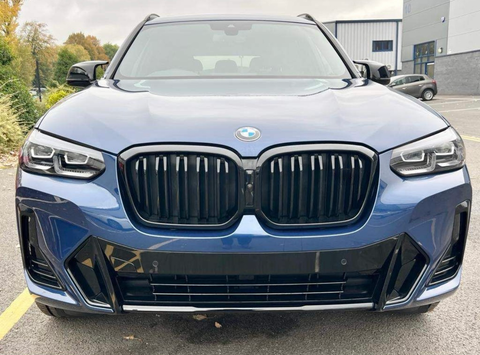 A blue BMW SUV is parked in a parking lot, displaying a prominent front grille and headlights, with trees and industrial buildings in the background.