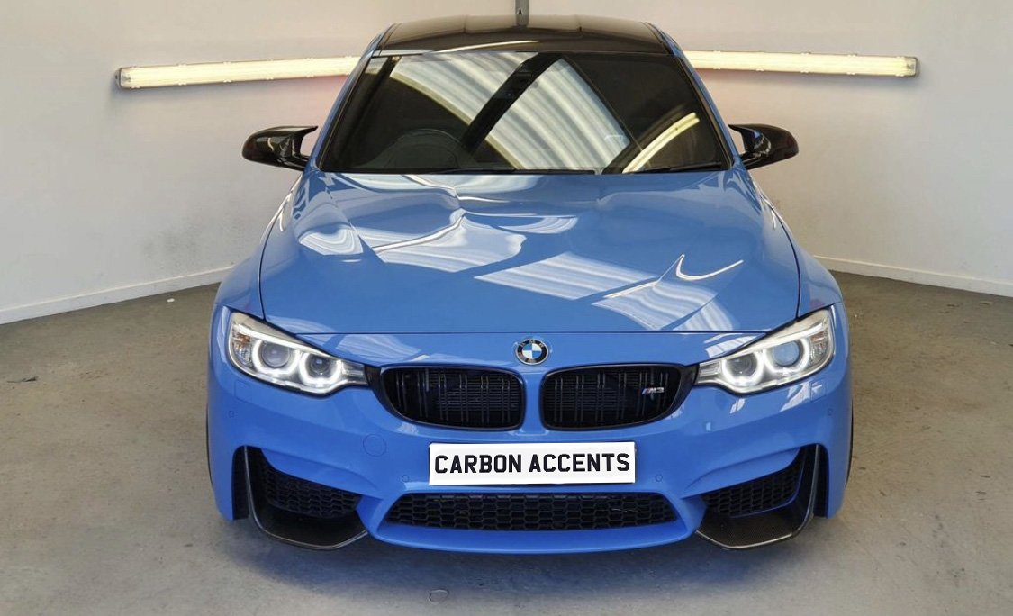 A blue BMW sports car, parked in a well-lit showroom, features the text "CARBON ACCENTS" on the license plate.
