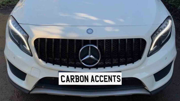 White car front with illuminated headlights and a distinct grille displaying a Mercedes logo. License plate reads "CARBON ACCENTS." The background is blurred with trees visible.