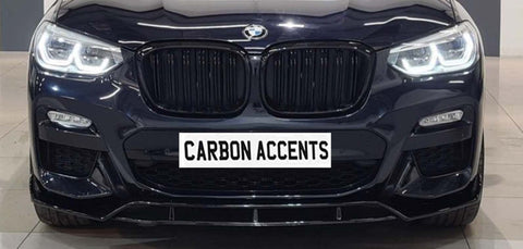Car front view displaying illuminated headlights in a showroom setting. Prominently featured below the grille is a sign reading "CARBON ACCENTS."
