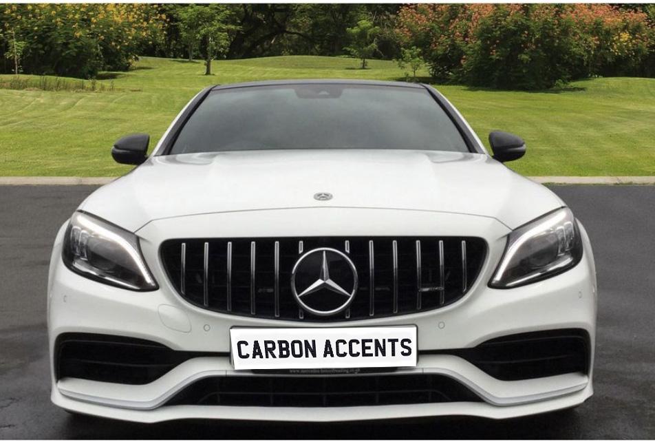A white car is parked on a driveway, displaying the text "CARBON ACCENTS" on its front plate. The background features a garden with grass and trees.