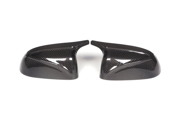 Two carbon fiber car mirror covers are placed side by side, angled inward, on a plain white background. The glossy surface reflects light, highlighting their textured pattern.