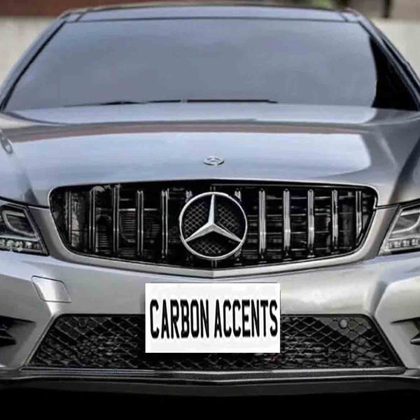 A silver car features a front grille with a central logo, parked in an urban area. The license plate reads "CARBON ACCENTS."