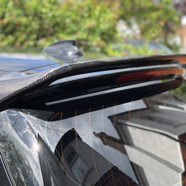 A carbon fiber car spoiler is attached to the rear of a black vehicle, glistening in sunlight. In the background, residential houses and greenery are slightly blurred.