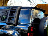 A black off-road vehicle with a mounted ladder and spare tire, reflecting trees and sky, is parked near a tree. A logo reads 