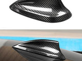 A carbon fiber shark fin antenna rests on a car roof, reflecting light. The surface around it shows a sky-blue hue, suggesting outdoor daylight conditions.