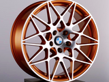 A bronze alloy wheel with intricate spoke design and a central blue and white emblem, resting on a black pedestal against a neutral gray background.